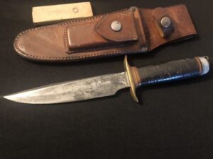 A vintage Bowie knife with a leather sheath on a wooden surface.