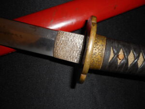 Close-up of a katana's handle and guard with a red scabbard.