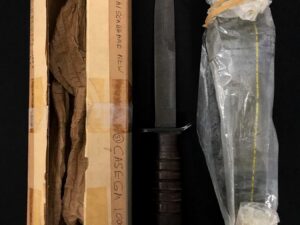 A knife with a black handle and sheath, next to a wooden box and a plastic-wrapped sheath.